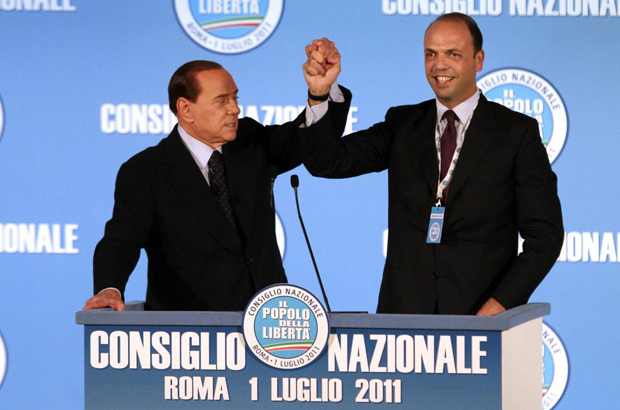 Silvio Berlusconi ed Angelino Alfano si stringono la mano sul palco di un comizio del Popolo delle libertà. Sul poggio c'è la scritta Consiglio Nazionale Roma 1 luglio 2011 ed il logo del partito