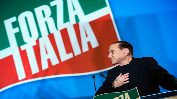 berlusconi che si tocca il cuore con dietro la bandiera di Fi