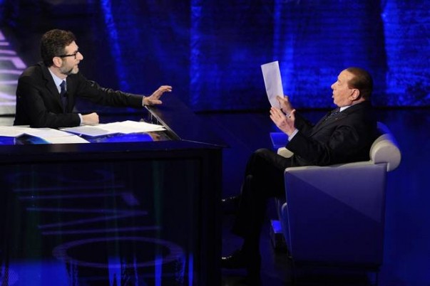 Silvio Berlusconi viene intervistato nello studio di che tempo che fa da Fabio Fazio. Fabio Fazio è seduto davanti ad una scrivania con la mano protesa verso Berlusconi. Berlusconi è seduto in poltrona e regge dei fogli.