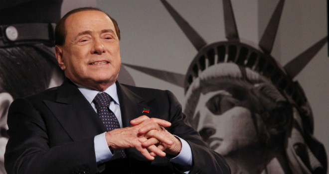 silbio berlusconi sorridente con le mani nelle mani e sullo sfondo la statua della liberta