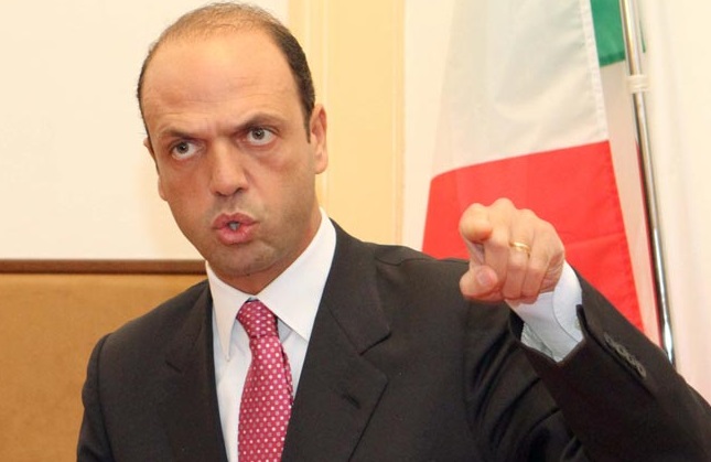 alfano con cravatta rosa e mano sinistra alta con indice alzato su sfondo bandiera italiana